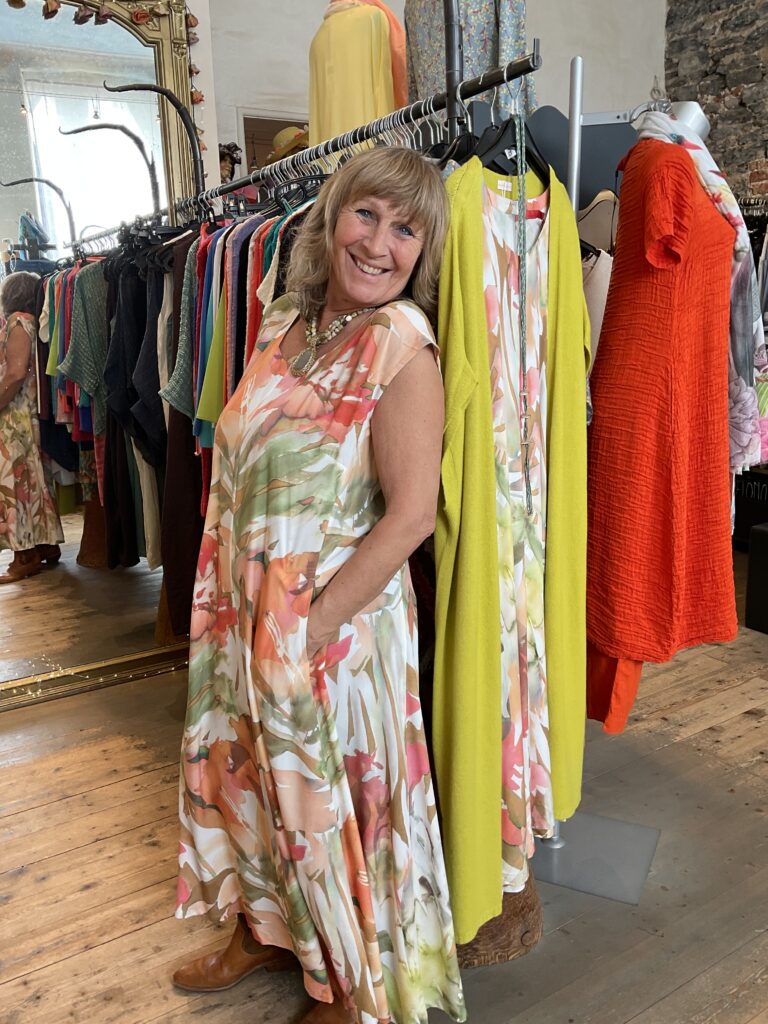 Kleurrijke, creatieve en unieke mode voor dames in hartje Gent. Grote maten mode dames kledij en accessoires voor vrouwen met een maatje meer of minder. Lange jurk met V-hals en korte kapmouw en florale print diverse kleuren ecru, groen, oranje, rood.